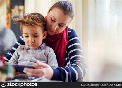Portrait of small caucasian boy little child sitting in lap of his mother female woman by the table at home or cafe restaurant in day front view mobile smart phone playing games learning teaching app