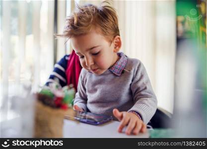 Portrait of small caucasian boy little child sitting in lap of his mother female woman by the table at home or cafe restaurant in day side view mobile smart phone playing games learning teaching app
