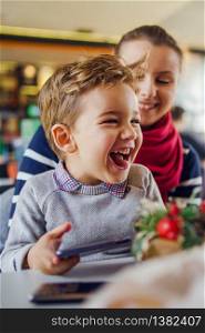 Portrait of small caucasian boy little child sitting in lap of his mother female woman by the table at home or cafe restaurant in day smiling front view happy family holding mobile smart phone