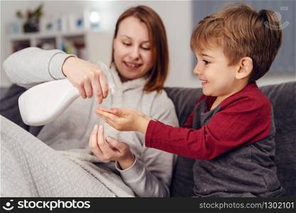 Portrait of small caucasian boy male child kid sitting on the sofa bed at home by his mother young woman spraying alcohol disinfectant antibacterial prevention hygiene product to his hands cleaning