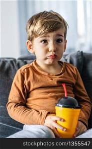 Portrait of small cute caucasian boy three or four years old sitting on the bed or sofa at home holding a plastic cup with straw drinking juice soda at home alone in day looking to camera half length