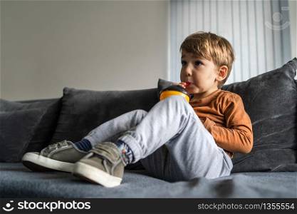 Portrait of small cute caucasian boy three or four years old sitting on the bed or sofa at home holding a plastic cup with straw drinking juice or soda at home alone in day looking to side full length