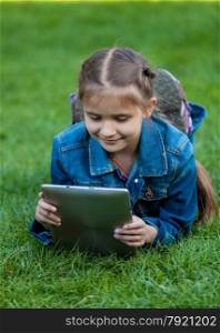 Portrait of small girl lying on grass and reading book from tablet