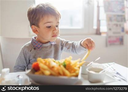 Portrait of small little cute caucasian boy kid eating french fries potato chips at the table in the restaurant or at home three or four years old