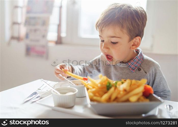Portrait of small little cute caucasian boy kid eating french fries potato chips at the table in the restaurant or at home three or four years old