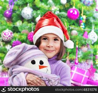 Portrait of small pretty girl wearing red santa hat hugging soft toy of white snowman over Christmas tree, New Year child's party