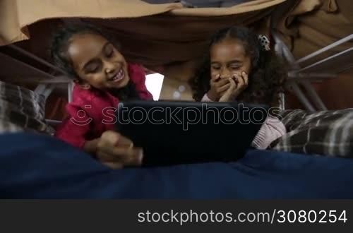 Portrait of smart african american kids using digital tablet pc in domestic interior. Adorable mixed race little girls playing online games on touchpad while lying in cubby house made of blanket and chairs. Closeup. Low view.