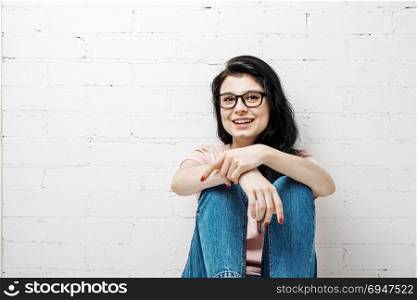 Portrait of smart beautiful brunette girl in eyeglasses with natural make-up sitting near white brick wall.. Portrait of smart beautiful brunette girl in eyeglasses with natural make-up sitting near white brick wall