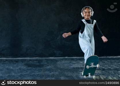 Portrait of smiling African American little kid boy with skateboard on black background. Happy adorable child wear white headphones, bib jeans, necklace chain in hip hop style