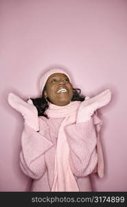 Portrait of smiling African-American mid-adult woman on pink background wearing winter coat, hat, and scarf looking upward.