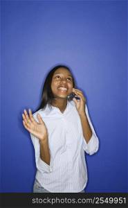 Portrait of smiling African-American teen girl looking up and talking on cellphone standing against blue background.