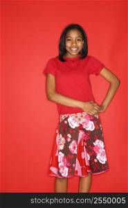 Portrait of smiling African-American teen girl with hands on her hips standing in front of red background.