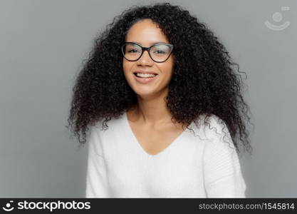 Portrait of smiling African American woman wears transparent eyewear and white jumper, being in high spirit, expresses good emotions, isolated over grey background. People and ethnicity concept