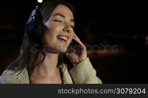 Portrait of smiling attractive girl with headphones listening to music while spending great time on summer night in the city. Gorgeous brunette female in earphones enjoying music at night outdoors. Slow motion. Steadicam stabilized shot.
