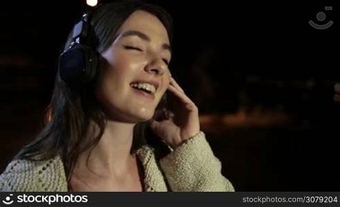 Portrait of smiling attractive girl with headphones listening to music while spending great time on summer night in the city. Gorgeous brunette female in earphones enjoying music at night outdoors. Slow motion. Steadicam stabilized shot.