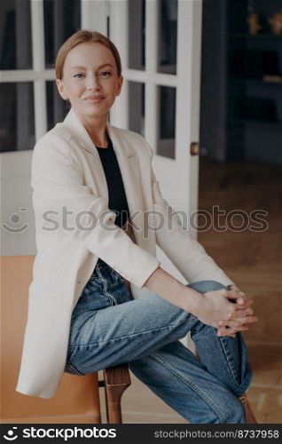 Portrait of smiling attractive young businesswoman wearing casual office clothing, sitting indoors. Confident pretty female entrepreneur in white suit jacket posing for photo looking at camera.. Smiling pretty businesswoman wearing casual office clothing, posing for portrait, looking at camera