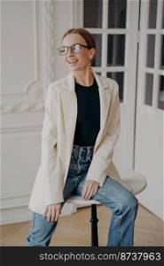 Portrait of smiling beautiful businesswoman sitting on little round table, posing for photo. Happy confident female fashion model wearing casual clothes, eyewear having fun, looking aside.. Happy pretty businesswoman in glasses sitting on little round table, posing for photo, looking aside
