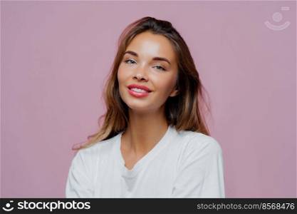 Portrait of smiling beautiful European woman with long hair, makeup, wears white casual jumper, feels delighted, poses at camera against purple background. People, beauty, skin care concept.