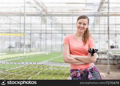 Portrait of smiling beautiful female botanist standing with arms crossed against herbs in greenhouse