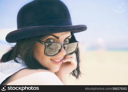 Portrait of smiling beautiful young woman in black hat. Model shooting on the beach.. Portrait of smiling beautiful young woman in black hat and retro sunglasses on the beach