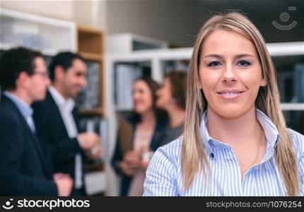 Portrait of smiling blonde woman looking at camera in the office with colleagues talking on background. Portrait of blonde woman looking at camera in office