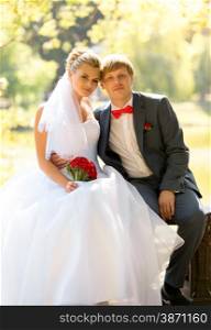 Portrait of smiling bride and groom sitting on bench at river under big tree