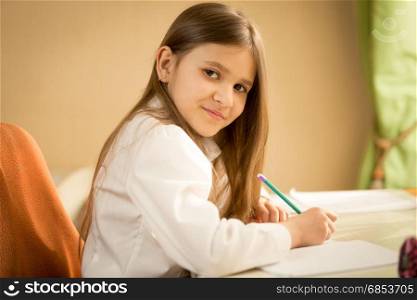 Portrait of smiling brunette girl in white shirt sitting behind desk and doing homework