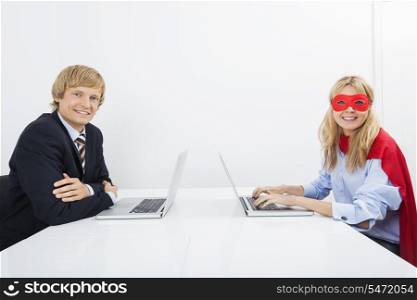 Portrait of smiling businessman with coworker in superhero costume using laptops at office