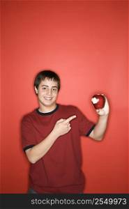 Portrait of smiling Caucasian teen boy pointing to partially eaten apple standing against red background.