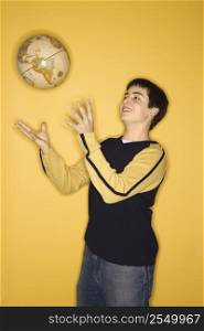 Portrait of smiling Caucasian teen boy tossing globe in air standing against yellow background.