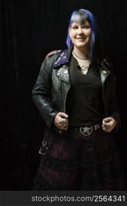 Portrait of smiling Caucasian woman with blue hair and black leather jacket standing against black background.