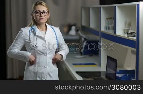 Portrait of smiling confident blonde female doctor with stethoscope wearing eyeglasses and white uniform standing over medical office background. Confident beautiful woman medic posing at hospital and looking at camera with joyful smile.