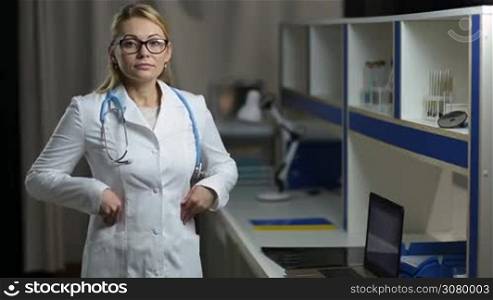 Portrait of smiling confident blonde female doctor with stethoscope wearing eyeglasses and white uniform standing over medical office background. Confident beautiful woman medic posing at hospital and looking at camera with joyful smile.