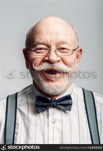 Portrait of smiling elderly man in a bow tie and glasses, grey background. Mature senior looking at camera in studio. Smiling elderly man in a bow tie and glasses