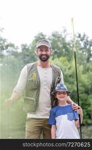 Portrait of smiling father and son standing with fishing rods against trees