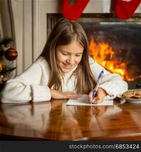 Portrait of smiling girl in sweater sitting by the fireplace and writing letter to Santa Claus