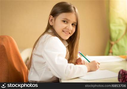 Portrait of smiling girl in white shirt sitting behind desk and doing homework
