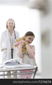 Portrait of smiling girl with teddybear standing against doctor at hospital