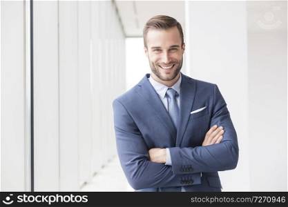Portrait of smiling handsome young businessman standing with arms crossed in new office