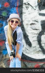 Portrait of smiling hipster girl with retro photo camera against urban wall outdoors