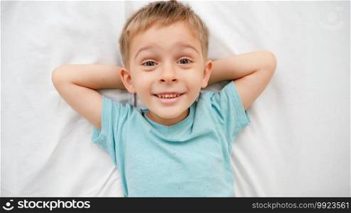 Portrait of smiling little boy in pajamas lying on big bed with white sheets and looking up in the camera. Concept of happy children having good time at home.. Portrait of smiling little boy in pajamas lying on big bed with white sheets and looking up in the camera. Concept of happy children having good time at home