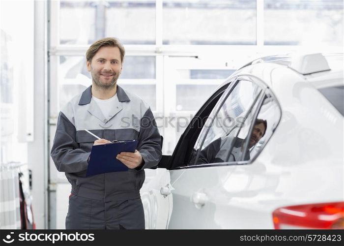 Portrait of smiling male automobile mechanic holding clipboard while standing by car in repair shop