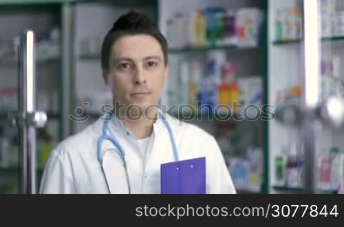Portrait of smiling male pharmacist in white coat standing over drugstore background. Cheerful pharmacist chemist man wearing stethoscope, holding clipboard in hand and looking at camera with toothy smile. Health care and pharmacology concept