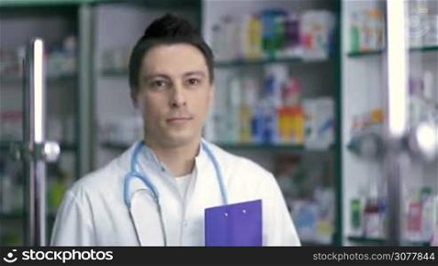 Portrait of smiling male pharmacist in white coat standing over drugstore background. Cheerful pharmacist chemist man wearing stethoscope, holding clipboard in hand and looking at camera with toothy smile. Health care and pharmacology concept