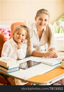 Portrait of smiling mother and daughter sitting at desk and looking at camera