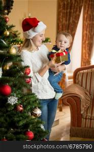 Portrait of smiling mother holding her baby boy at Christmas tree and giving him present