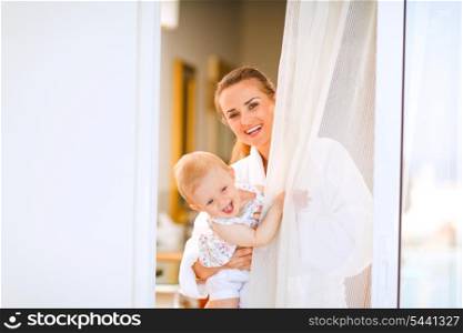 Portrait of smiling mother in bathrobe with baby looking out from window&#xA;