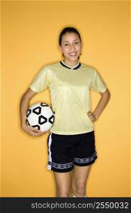 Portrait of smiling Multi-racial teen girl holding soccer ball at hip standing against yellow background.