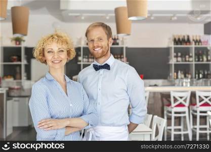 Portrait of smiling owner and waiter standing against furniture at restaurant