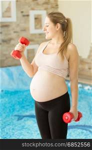Portrait of smiling pregnant woman posing with dumbbells at gym